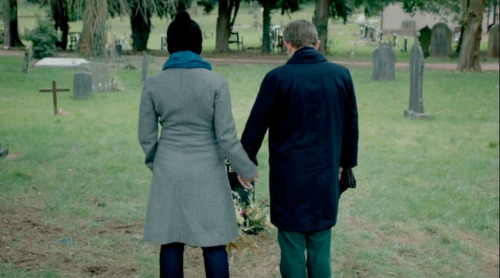 So I honestly can&#8217;t believe I&#8217;ve never seen anyone talking about the art direction of this scene. If I&#8217;m repeating something, Ah,well. But I&#8217;ve honestly never seen it pointed out that this is the very first time we see Mary, and there are three important things here:
Mary reaches for John&#8217;s hand. John takes it, of course&#8212;he is used to being offered comfort for his loss, by now&#8212;but he is not reaching out to her for comfort in his sadness. She is inserting herself into his grief. Reflexively, he lets her.
We only see the back of her. It&#8217;s unusual to introduce a major protagonist any other way than by showing their face pretty much immediately. A major antagonist, however&#8230;a baddie&#8230;well, they often are introduced in a cloud of cigarette smoke, from a distance, in the shadows, as a mysterious voice on a phone, or in some other way that doesn&#8217;t tell us right away who they are. Our first glimpse of Mary gives us only the most vague information about her. Obviously a woman, obviously someone John is close to, as he holds her hand. Other than that&#8230;who is she? We don&#8217;t know.
Finally, it&#8217;s no mistake she is wearing a long, grey coat which flares slightly from the waist, and a blue scarf. But they are paler shades of those colours than Sherlock&#8217;s coat and scarf were, because Mary is but a pale imitation of the person we are used to seeing standing beside John Watson (even once, when they were handcuffed together, holding John Watson&#8217;s hand in a manner similar to what we see here). Her coat and scarf look cheap, &#8220;less than,&#8221; and her denim jeans are &#8220;less&#8221; than Sherlock Holmes&#8217;s designer trousers. Her dark hat is a visual echo of Sherlock&#8217;s dark hair. This whole shot is set up not only to remind us that Sherlock used to stand here at John Watson&#8217;s side, but also that This is some lesser, fake, replacement-Sherlock standing at John Watson&#8217;s side, and whether consciously or unconsciously, John has chosen a pale imitation indeed.