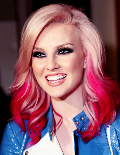 Perrie Edwards smiling and looking off in the distance. She has fake eyelashes on and a pink lip. Her hair is pink ombre and she is wearing a blue jacket.