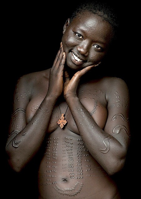 Native Nudity (nativenudity): By Eric Lafforgue She is called Anna, she lives in Hana Mursi village, in Omo valley, Ethiopia. She is from Bodi tribe. She had all those scarifications when she was 14. Since last year she has became christian, so she wears the cross around her neck and most of the time a tshirt, apart when she has to cross the river to come back to her village.. She is the symbol of all the tribes i meet and who are changing so quick…for many different reasons. Her village has only 300 people, the government has settled a giant sugarcane factory, and plans to bring 100 000 people from all over the country in the next years. I took this picture just at the entrance of her house, with natural light. everything is simple and works so good when the “model” is wonderful.